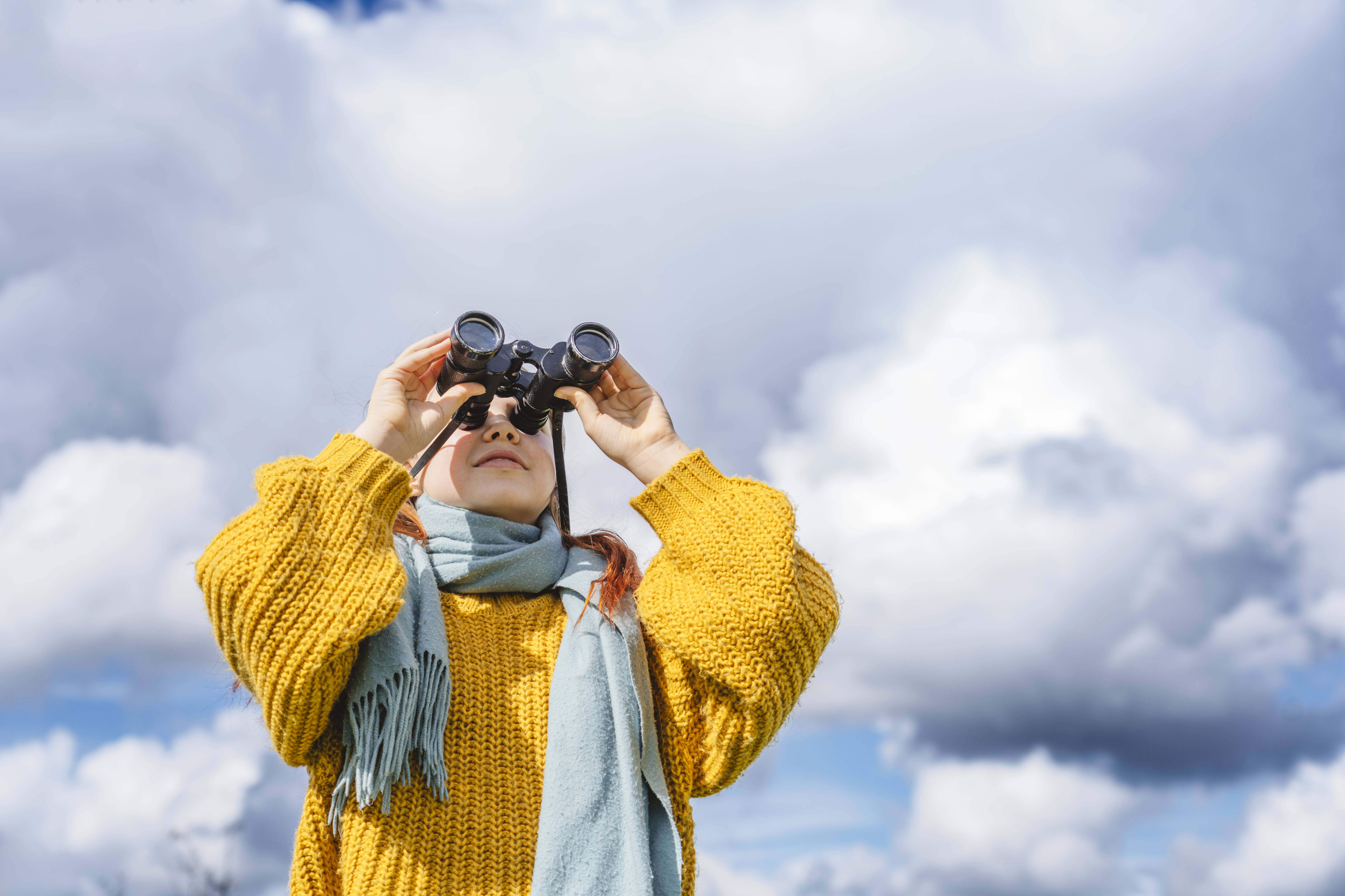 Blick aufs Soziale Eine Frau in einer gelben Strickjacke vor blauem Himmel mit weißen Wolken, sie blickt durch ein Fernglas.