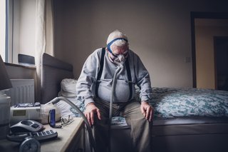 Älterer Mann sitzt zu Hause auf seinem Bett. Er trägt ein medizinisches Beatmungsgerät über seinem Gesicht, das an einer Maschine neben ihm befestigt ist