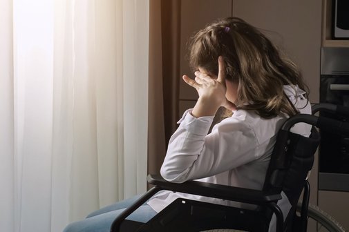 Frau mit einer Behinderung im Rollstuhl Eine junge Frau mit Behinderung sitzt in einem Rollstuhl an einem Fenster. Sie blickt nachdenklich oder sehnsüchtig nach draußen, während warmes Sonnenlicht durch das Fenster fällt und sie sowie den Vorhang im Hintergrund beleuchtet.