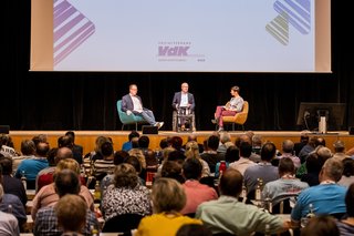 Blick auf die Bühne der SBV-Konferenz: Die Personen sitzen auf der Bühne und diskutieren, im Vordergrund sind die Besucher zu sehen.