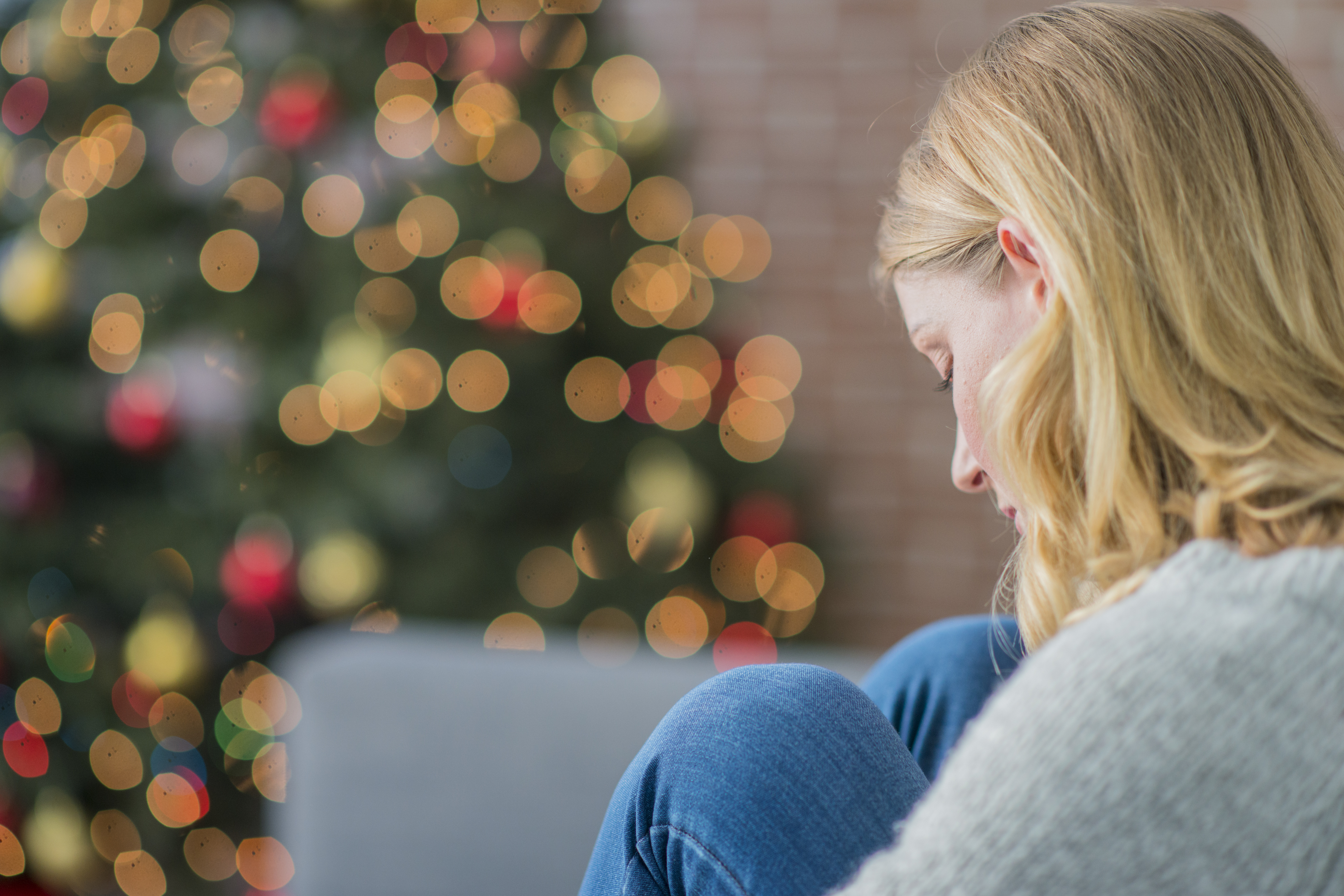 Einsamkeit Weihnachten Feiertage wie damit umgehen Allein zur Weihnachtszeit: Junge Fraue sitzt auf ihrem Sofa, die Beine angewinkelt und mit den Händen umklammert. Sie starrt traurig nach unten, im Hintergrund sind die Lichter eines Weihnachtsbaumes zu sehen.