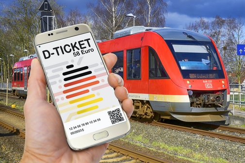 In einer Hand sieht man ein Smartphone. Auf dem Bildschirm ist das Deutschlandticket abgebildet. Im Bild rechts hinten sieht man einen Zug der Deutschen Bahn auf einem Gleis stehen.