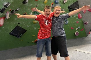 Das Foto zeigt Kinder mit Down-Syndrom in einer Boulderhalle. Sie halten sich in den Armen und lächeln in die Kamera. 