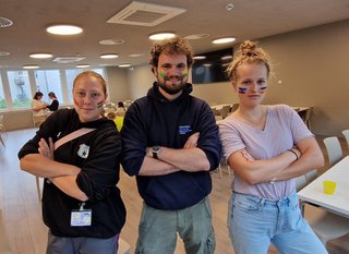 Auf dem Foto sieht man zwei junge Frauen und einen Mann nebeneinander stehen. Sie haben die Arme verschränkt und schauen in die Kamera. 