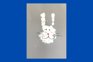 Das Bild zeigt ein weißes Hasenköpfchen, das durch eine bemalte Hand auf ein Papier gebracht wurde.