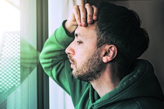 Ein Mann lehnt sich mit einem Arm an ein Fenster und fasst sich dabei an die Stirn.