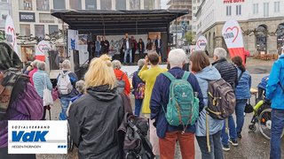 Das Bild zeigt die Bühne der Veranstaltung in Kassel mit einer guten Anzahl von Zuschauern. Sie tragen durchnässte Regenkleidung.
