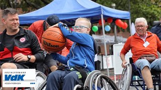 Auf dem Bild sieht man ein Rollstuhl-Basketball-Feld. Im MIttelpunkt setzt ein kleiner Junge im Rollstuhl zum Korbwurf an. Er zeigt viel Freude.
