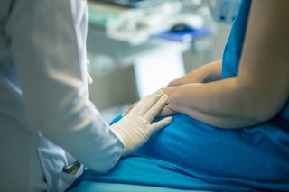 Foto einer Patientin in blauem Kittel und eine Ärztin, die ihre Hand auf ihre Hand legt