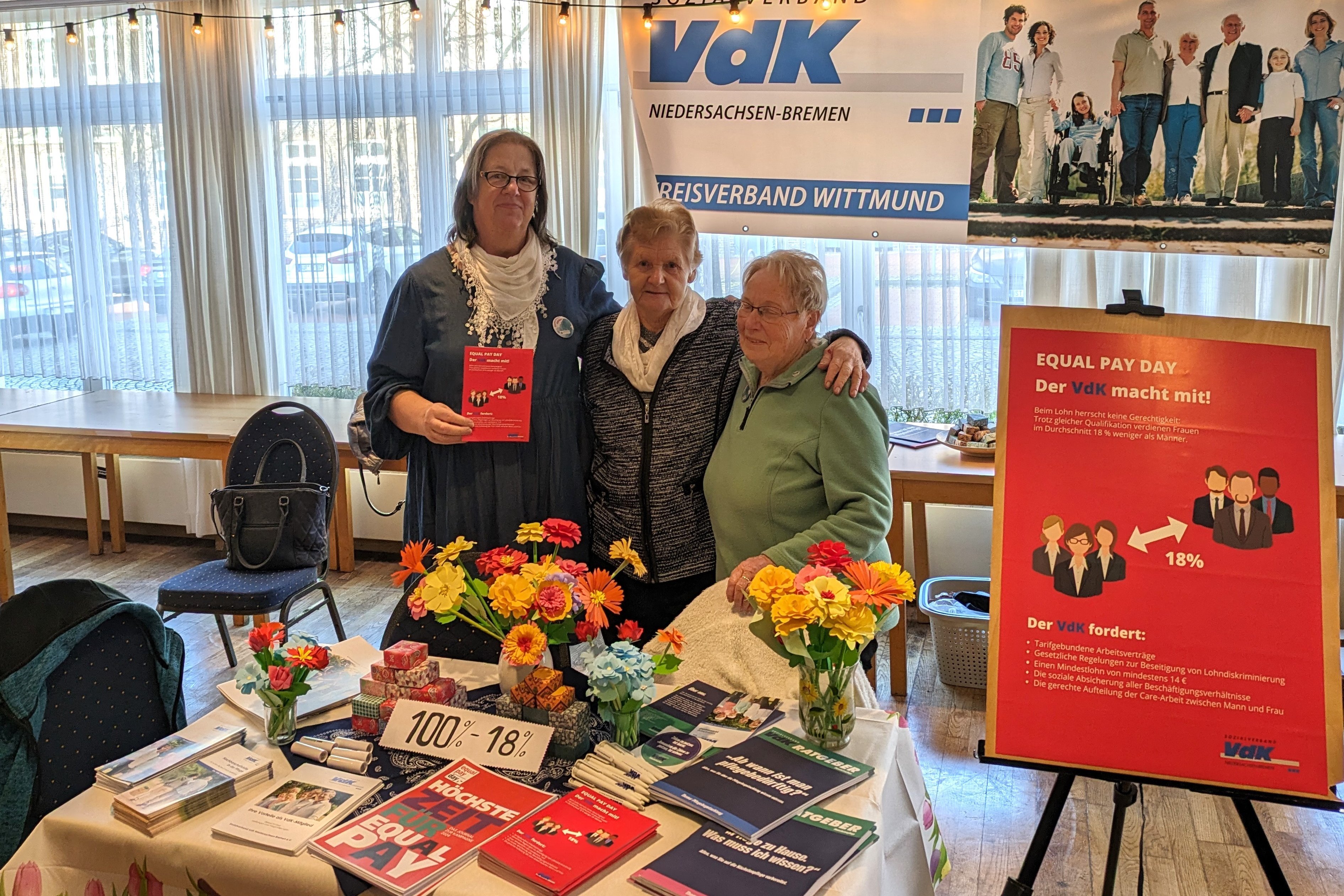 Drei Frauen an ihrem bunten Infostand neben einem roten Plakat zum Equal Pay Day