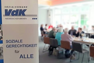 Ein blau-weißer VdK-Banner mit Logo und im Hintergrund Menschen an einem Tisch