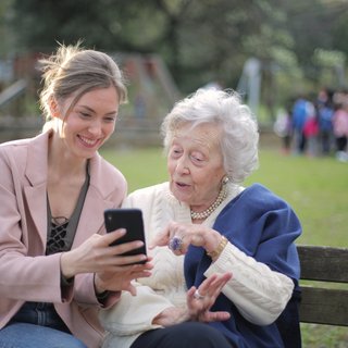 Eine jüngere Frau zeigt einer älteren Frau etwas auf dem Smartphone. Sie sitzen auf einer Bank im Park.