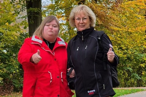 Zwei Frauen stehen auf einem Parkweg. Die Frau links tr&auml;gt eine rote Jacke, braune Hose und schwarze Schuhe. Die Frau rechts tr&auml;gt eine schwarze Jacke, rote Hose und graue Stiefeletten. Beide zeigen mit einer Hand den Daumen nach oben und l&auml;cheln. Im Hintergrund herbstlich gef&auml;rbte B&auml;ume mit gelb-gr&uuml;nem Laub.