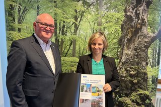 VdK-Landesverbandsvorsitzender und Mobilitätsministerin stehen gemeinsam mit VdK-Fotoband vor einem Plakat