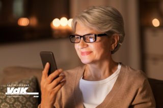 Ältere Frau mit Brille sitzt auf dem Sofa und schaut zufrieden auf ihr Smartphone.