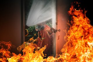 Ein brennender Hauseingang. Die Tür ist geöffnet. Zu sehen ist ein Feuerwehrmann und ein Schlauch, der das Feuer löscht.