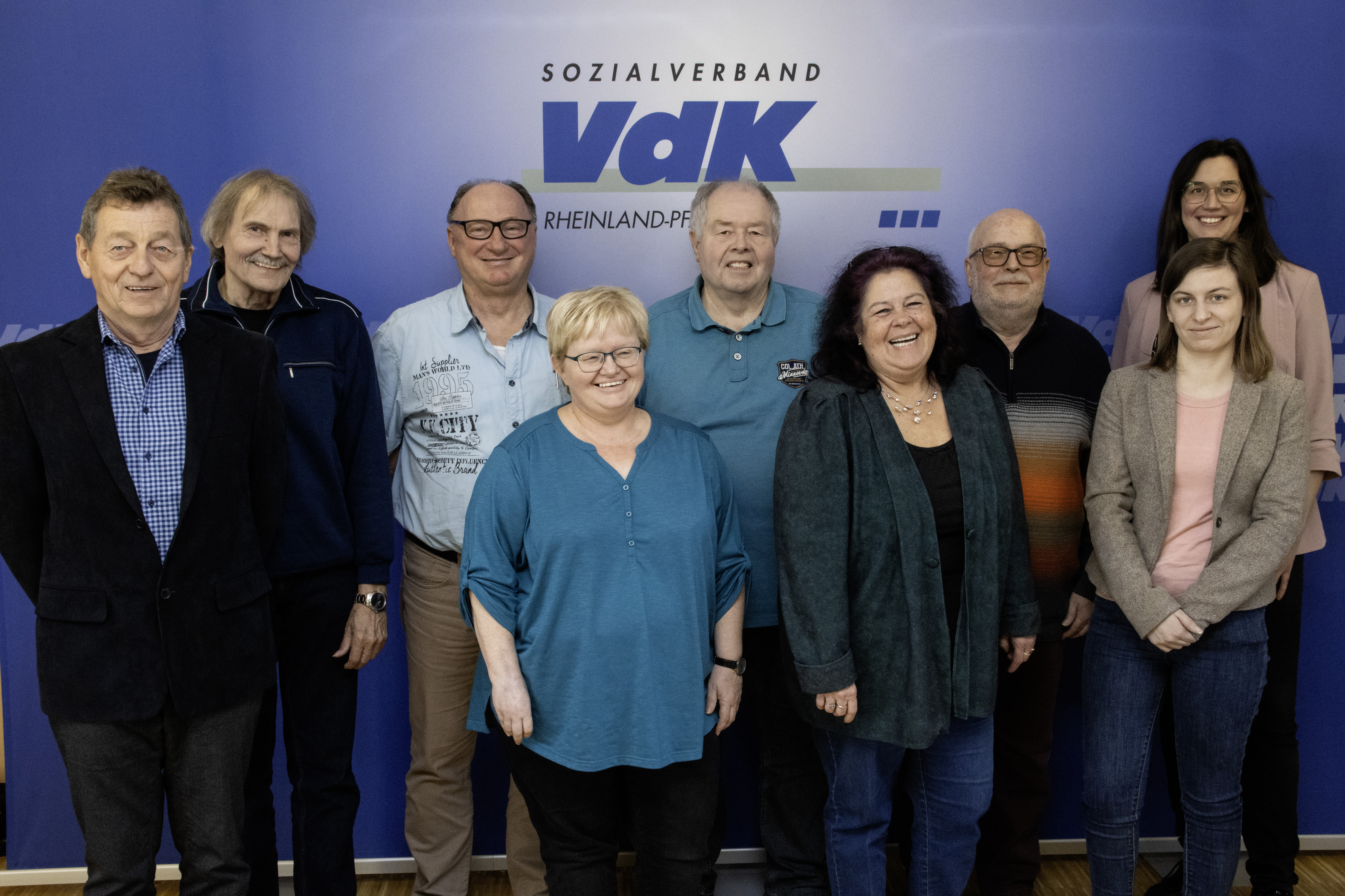 Frauen und Männer stehen als Gruppe vor einem blauen Hintergrund mit VdK-Logo.