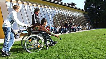 ein Junge schiebt einen anderen Jungen mit einem Rollstuhl über eine Wiese