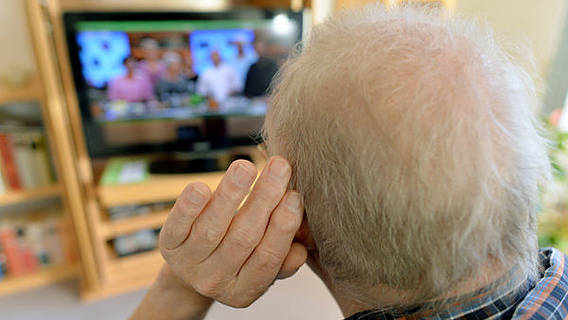 Symbolfoto: Ein älterer Herr sitzt vor dem Fernseher und hält sich horchend eine Hand vors Ohr.