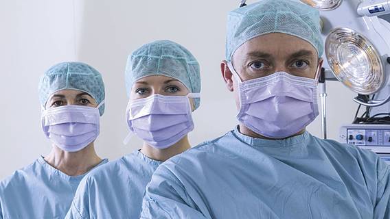 Symbolfoto: Drei Chirurginnen und Chirurgen in OP-Kleidung in einem OP-Saal. Sie tragen Mundschutz, OP-Haube und OP-Kittel.