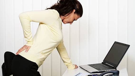 Symbolfoto: Junge Frau steht am Schreibstisch, fasst sich schmerzerfüllt an den Rücken