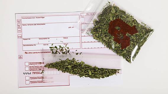 Symbolfoto: Ein leeres Rezept vom Arzt, darauf liegt ein mit Cannabisblättern gefülltes Tütchen mit einem Apotheken-Symbol darauf
