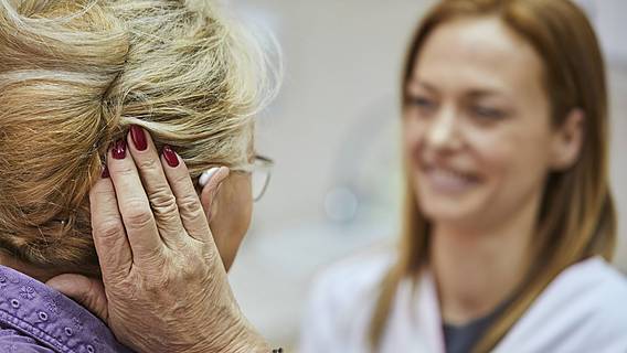 Symbolfoto: Ältere Frau probiert ein Hörgerät an, hält sich eine Hand ans Ohr. Vor ihr steht eine jüngere Frau im weißen Kittel, vermutlich eine Ärztin oder Hörgeräteakustikerin