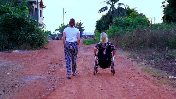 Zwei Frauen von hinten auf einem sandigen Weg, eine im Rollstuhl, die andere nicht