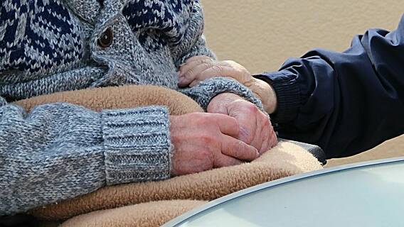 Eine ältere Person sitzt mit an einem Tisch, eine andere Person hat die Hand auf ihren Arm gelegt.