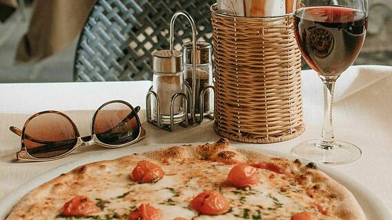Eine Pizza und ein Glas Rotwein auf einem Tisch, daneben eine Sonnenbrille