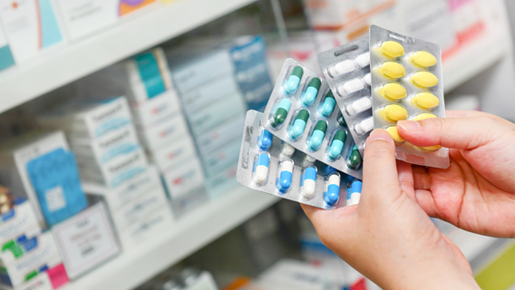 Das Bild zeigt zwei Hände, die viele Tabletten-Packungen halten. Im Hintergrund ist ein Regal mit Medikamenten zu sehen.