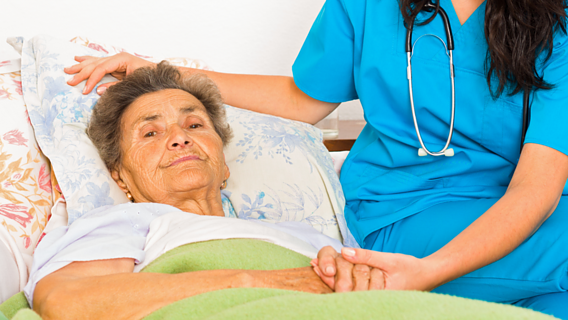 Eine ältere Frau liegt im Bett zu hause und eine Pflegekraft sitzt an ihrer Seite