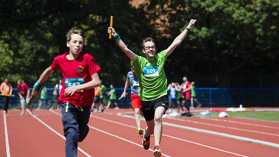 Teilnehmer bei den Special Olympics auf der Laufbahn - ein Läufer reißt die Arme hoch, hält einen Staffelstab. Ein anderer sprintet kurz vor ihm.