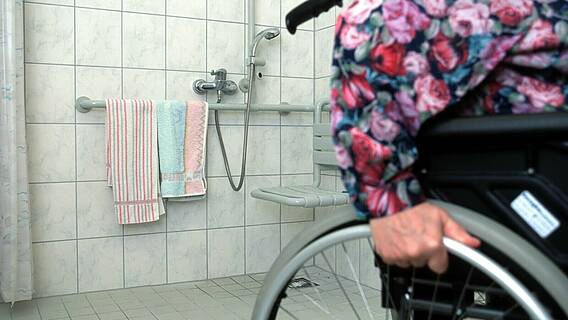 Symbolfoto: Eine Frau im Rollstuhl neben einer barrierefreien Dusche mit Duschsitz und ebenerdigem Einstieg