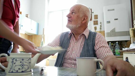 Symbolfoto: Älterer Mann sitzt am Küchentisch, eine Frau reicht ihm einen Teller mit Essen