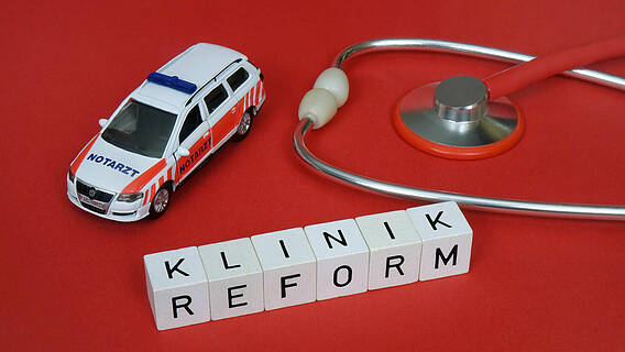 Symbolfoto: Neben dem Schriftzug Klinikreform, der aus Buchstabenwürfeln gebildet wird, befinden sich ein Spielzeug-Notarztfahrzeug und ein Stethoskop