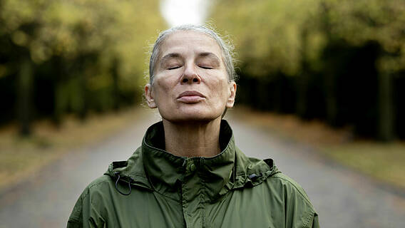 Symbolfoto: Ältere Frau mit grüner Regenjacke im Wald, sie hat die Augen geschlossen und atmet tief durch