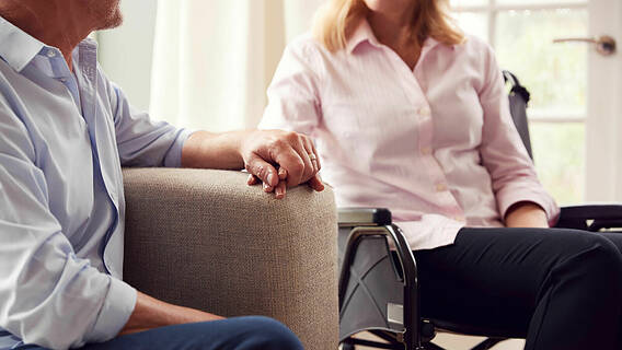 Symbolfoto: Älterer Mann auf einem Sofa und ältere Frau im Rollstuhl sitzen vertraut nebeneinander, halten sich an der Hand.