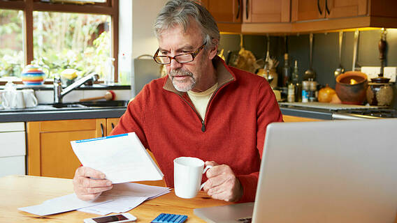 Symbolfoto: Älterer Mann sitzt in seiner Küche am Tisch, hat eine Kaffeetasse in der Hand und schaut Unterlagen an. Auf dem Tisch steht ein Notebook und ein Taschenrechner liegt vor dem Mann.