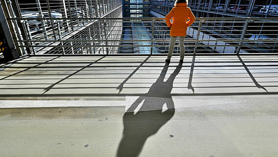 Symbolfoto: Eine Frau steht an einem Geländer, die Sonne steht so, dass ihr Schatten sehr lang auf den Boden fällt.