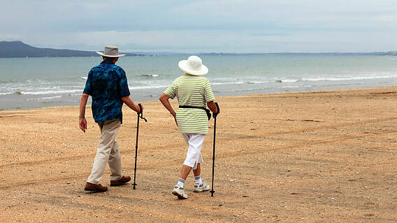 Symbolfoto: Älteres Paar geht am Strand spazieren, beide tragen Sonnenhüte und haben einen Gehstock