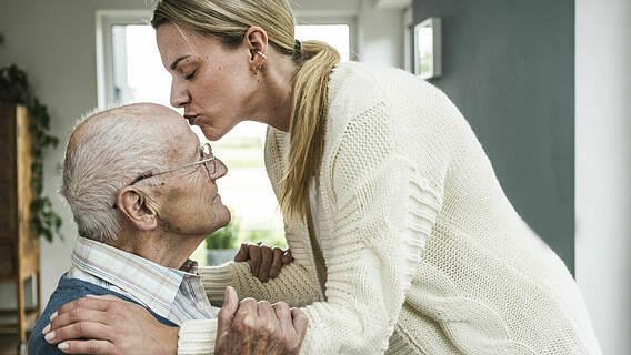 Symbolfoto: Jüngere Frau küsst älteren Mann liebevoll auf die Stirn, dabei steht sie und er sitzt, sie beugt sich zu ihm herunter.