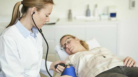 Symbolfoto: Eine Ärztin misst bei einer jungen Frau, die auf einer Untersuchungsliege liegt, den Blutdruck