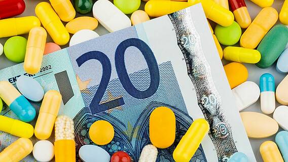 Symbolfoto für Kosten von Medikamenten und Krankenkasse: Ein 20-Euro-Geldschein liegt auf einem Haufen bunter Tabletten