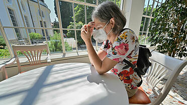 Symvolfoto: Eine Frau mit einer FFP2-Maske sitzt an einem Tisch, hat erschöpft den Kopf an die Hände gelegt
