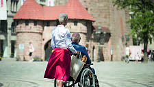 Das Bild zeigt eine ältere Frau, die einen älteren Mann im Rollstuhl schiebt.