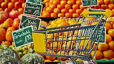 Ein Einkaufswagen, im Hintergrund Supermarkt-Auslagen mit Obst und Gemüse.