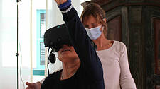 Eine Heimbewohnerin mit VR-Brille streckt einen Arm nach oben, hinter ihr steht die Sozialpädagogin Alexandra Kasper.