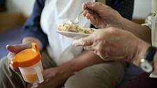 Symbolfoto: Hände eines älteren Mannes halten eine Schnabeltasse. Hände einer älteren Frau reichen Essen von einem Teller an.