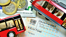 Ein 9-Euro-Ticket, Euro-Münzen und Scheine und zwei kleine Modell-Fahrzeige: eine Bahnm und ein Bus